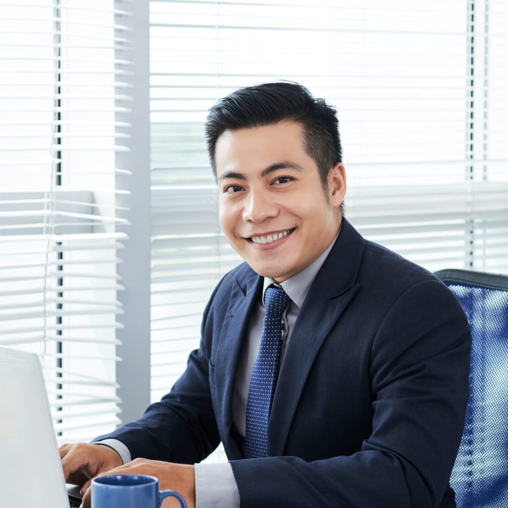 Waist-up portrait of handsome Asian businessman wearing classical suit posing for photography with wide smile while finishing ambitious project on laptop, modern office interior on background